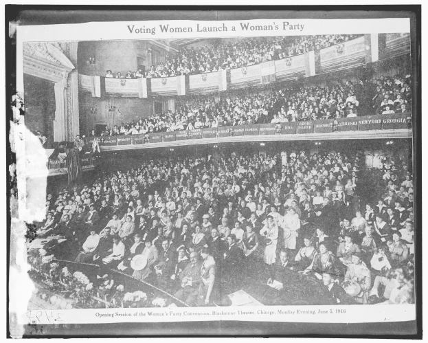 Ein Schwarz-Wei├č-Foto einer gro├čen Menge von Menschen in einem Geb├Ąude, einige sitzen, einige stehen, Schilder mit Text und eine Wand im Hintergrund. Unten steht der Text "Wahlberechtigte Frauen gr├╝nden eine Frauenpartei."