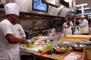 Eine Gruppe von Köchen in einer Küche, die Essen auf einem Schneidbrett zubereiten, mit einem Koch im Vordergrund in einer weißen Schürze und Mütze, der ein Messer hält, und umgeben von Utensilien, Schüsseln, Gemüse, einem Mobiltelefon, Flaschen und anderen Gegenständen, mit einem Fernseher und Deckenlampen im Hintergrund.