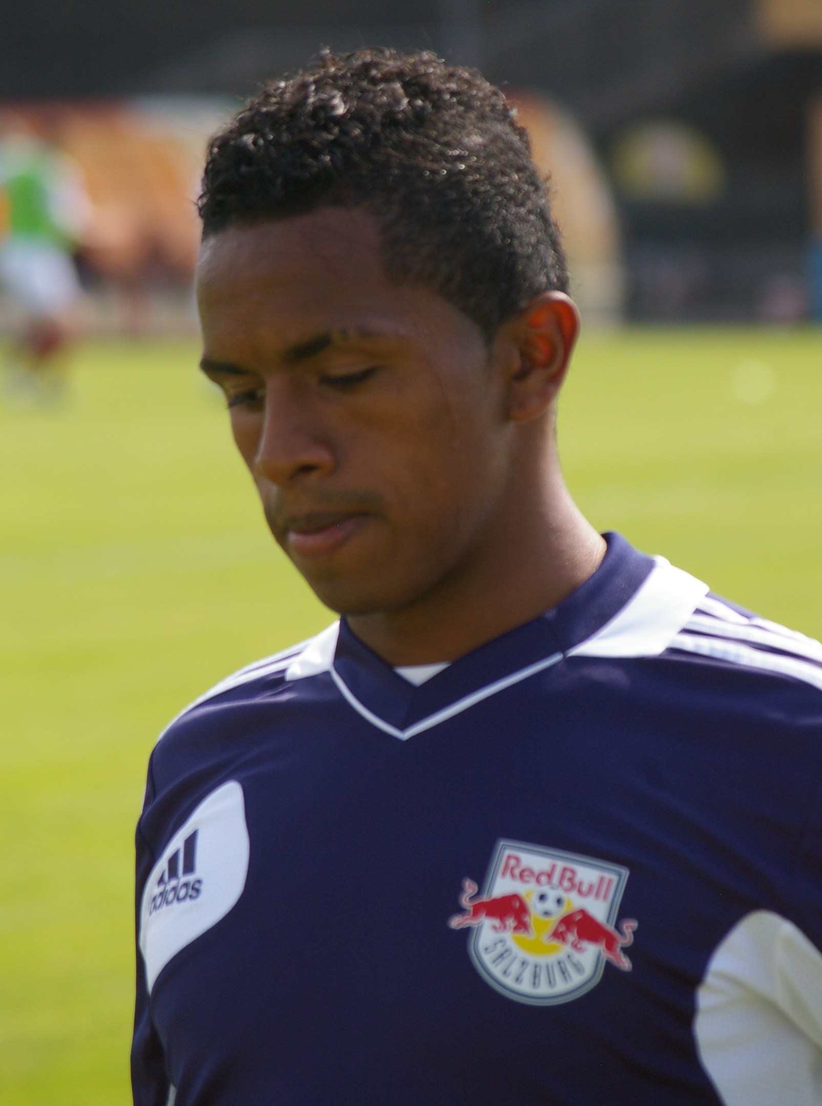Ein junger Mann in einem blauen T-Shirt steht auf einem Fußballfeld und sieht aus wie ein Flügelspieler mit kurzen dunklen Haaren und einem entschlossenen Gesichtsausdruck.