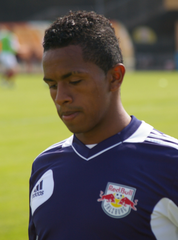 Ein junger Mann in einem blauen T-Shirt steht auf einem Fußballfeld und sieht aus wie ein Flügelspieler mit kurzen dunklen Haaren und einem entschlossenen Gesichtsausdruck.