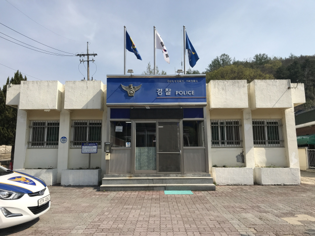 Eine Polizeistation mit einem geparkten Auto davor, das Fenster, eine Tür, Fahnen auf Stangen, ein Gebäude-Textschild, einen Strommast mit Drähten, bewölkten Himmel und entfernte Bäume zeigt.