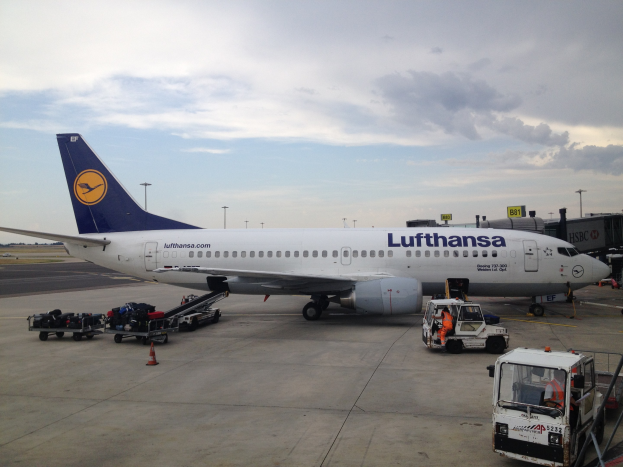 Ein Lufthansa Airbus A320-200 ist am Frankfurter Airport geparkt, mit verschiedenen Bodenfahrzeugen, Wagen, Verkehrskegeln, Pfählen und Texttafeln im Bild.