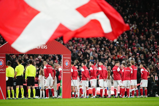 Gruppe von Menschen auf einem Fußballfeld mit einer roten und weißen Fahne im Vordergrund, einem Bogen mit der Aufschrift "Bayern München vs. Bayern München Betting Tips & Preview" im Hintergrund und einer großen Menge im Stadion.