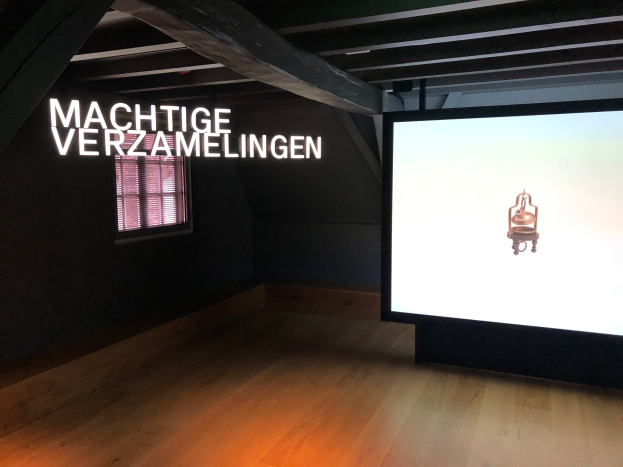 Installationsansicht der Ausstellung 'Machtige Sammlungen' im Museum of Modern Art in Berlin, mit einem großen Bildschirm mit Text an der Wand rechts und einem Fenster links.