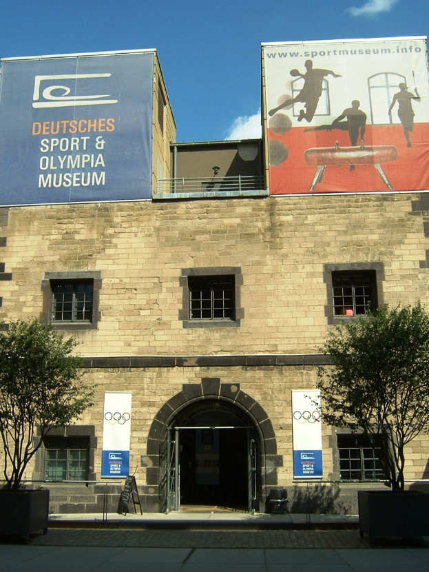 Außenansicht des Deutschen Sport & Olympiamuseums in Berlin, das ein Gebäude mit Fenstern und einer Tür zeigt, umgeben von Bäumen und Bannern, unter einem bewölkten Himmel.