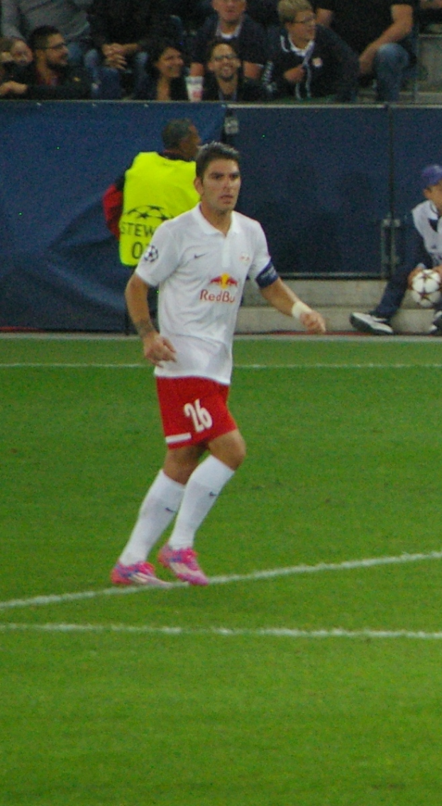 Ein Mann in einem weißen Hemd und roten Shorts, der auf einem Feld Füßball spielt, umgeben von einer Gruppe von Menschen im Hintergrund.