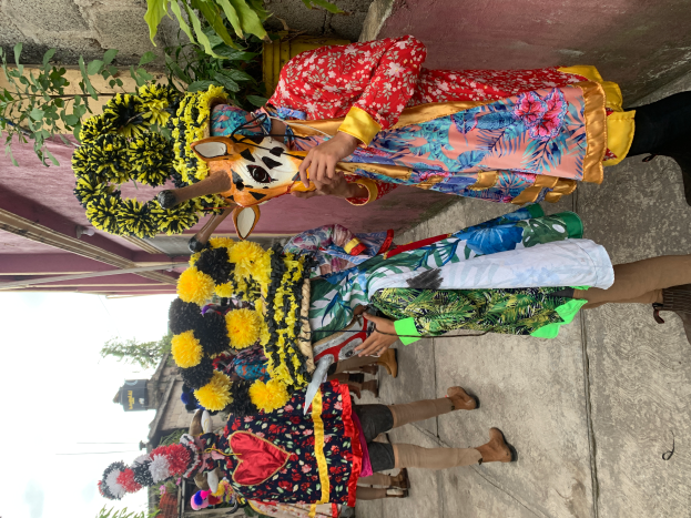 Eine Gruppe von Menschen in bunten Kostümen steht zusammen vor einem Gebäude, hält verschiedene Gegenstände, mit Bäumen, Gebäuden und einem klaren blauen Himmel im Hintergrund bei einem Karnevalsumzug.