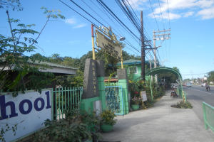 Einfahrt zu einer Schule mit einem "Schule"-Schild, Fahrzeuge auf der Straße, ein Tor, ein Tor, eine Namensschild, Pflanzen, Bäume, Strommasten mit Drähten und ein bewölkter Himmel.