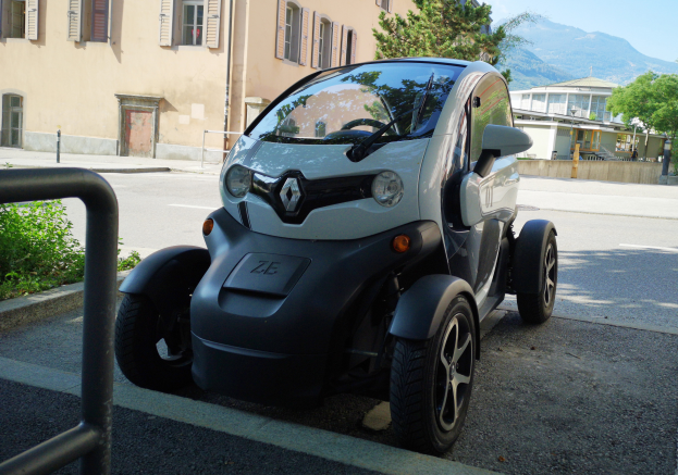 Ein Renault Twizy Elektroauto steht auf der Straße vor einem Gebäude mit Fenstern und Türen, umgeben von Pflanzen und einem Geländer, mit Bäumen, Gebäuden, Hügeln und einem klaren blauen Himmel im Hintergrund.