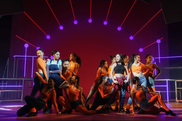 Eine Gruppe junger Tänzer in bunten Kostümen performs auf einer Bühne mit Geländern, beleuchtet von bunten Lichtern und umgeben von geometrischen Bühnen-Dekorationen, während ein applaudierendes Publikum zusieht.