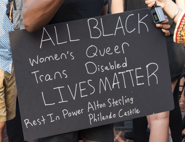 Eine Gruppe von Menschen, die zusammenstehen, wobei eine Person in der Mitte ein Schild hält, auf dem "All Black Women's Queer Trans-Disabled Lives Matter" steht, und eine weitere Person ein Objekt hält, sowie sichtbare Stühle im Hintergrund.