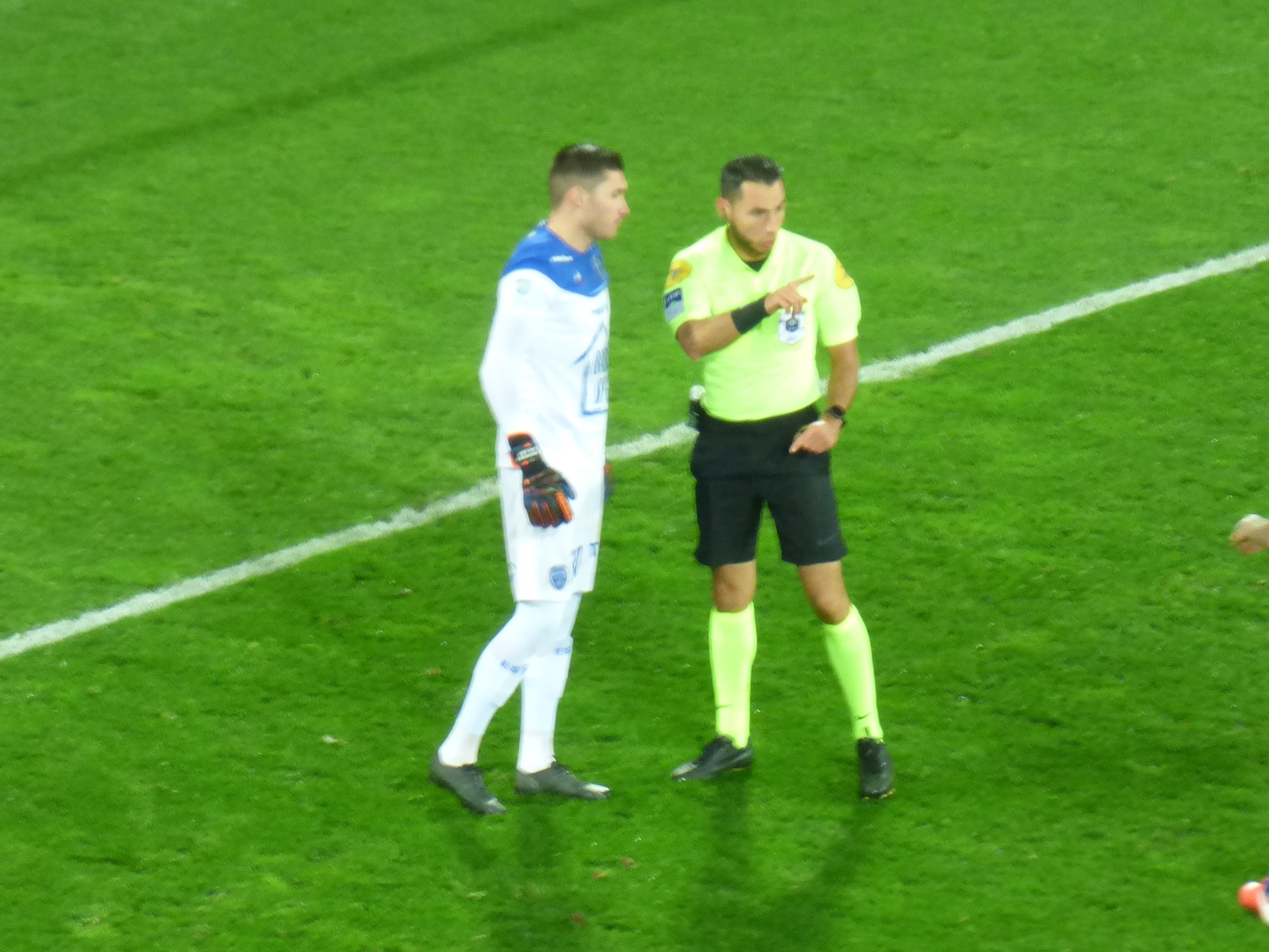 Schiedsrichter in gelbem T-Shirt und schwarzen Shorts spricht mit Fußballer in weiß-blauer Uniform auf dem Feld, beide tragen Turnschuhe.