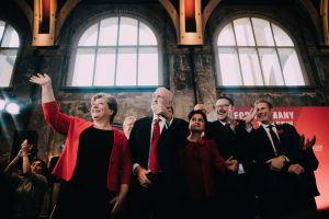 Eine Gruppe von Menschen, Mitglieder der Labour Party, stehen klatschend vor einer Menge, mit einem Banner, Glasfenstern und beleuchteten Lichtern im Hintergrund.