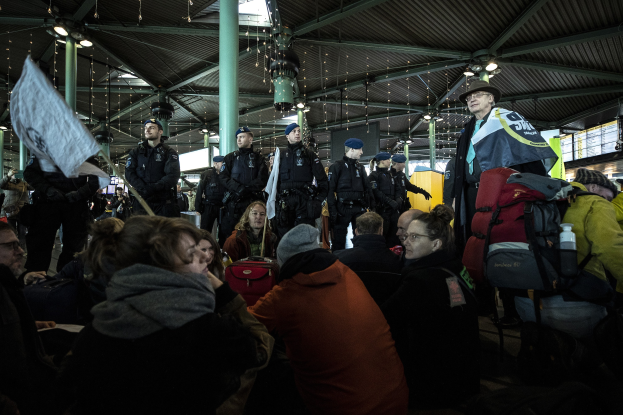 Eine Gruppe von Menschen steht vor einer Menschenmenge in einem Bahnhof, einige halten Schilder und Transparente, mit Säulen und Deckenlampen im Hintergrund.