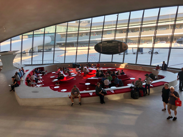 Eine große Gruppe von Menschen sitzt und steht in einem Flughafen-Terminal mit verstreuten Stühlen und Tischen, einer Treppe auf der linken Seite und Fahrzeugen, Gebäuden und einem klaren Himmel im Hintergrund.
