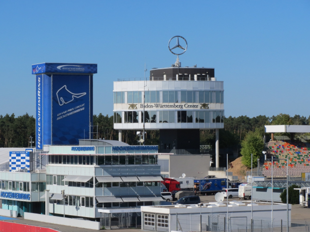 Außenansicht des Mercedes-AMG Petronas F1 Team Headquarters in Berlin, Deutschland, mit Gebäuden, einem Zaun, Fahrzeugen, Bäumen und einem Logo auf der Struktur.