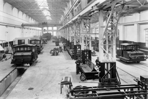 Ein Schwarz-Weiß-Foto einer Fabrikhalle mit Arbeitern, die an Zügen auf dem Boden arbeiten, umgeben von Metallstangen, Deckenlampen und Wandfenstern.