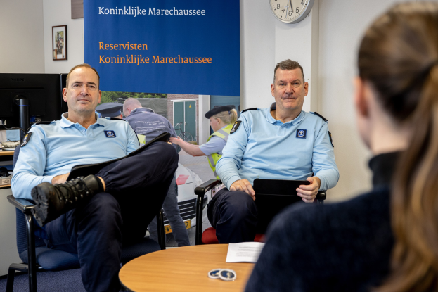 Zwei Polizeibeamte sitzen an einem Tisch mit Papieren, sprechen mit einer Frau; eine Wand mit einem Banner, einer Uhr und Fotorahmen im Hintergrund und ein weiterer Tisch mit einem Monitor, Tastatur und Maus links.