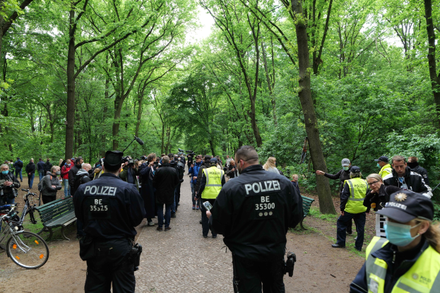 Eine Gruppe von Polizisten steht vor einer Menschenmenge, einige tragen Mützen und Masken, mit Fahrrädern und einer Bank im Vordergrund und Bäumen und Himmel im Hintergrund, wahrscheinlich während einer Anti-Terror-Demonstration in Berlin.