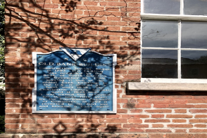Altes Bankgebäude mit blauer Tafel an der Seite, einem Fenster auf der rechten Seite und einem Baum auf der linken Seite.