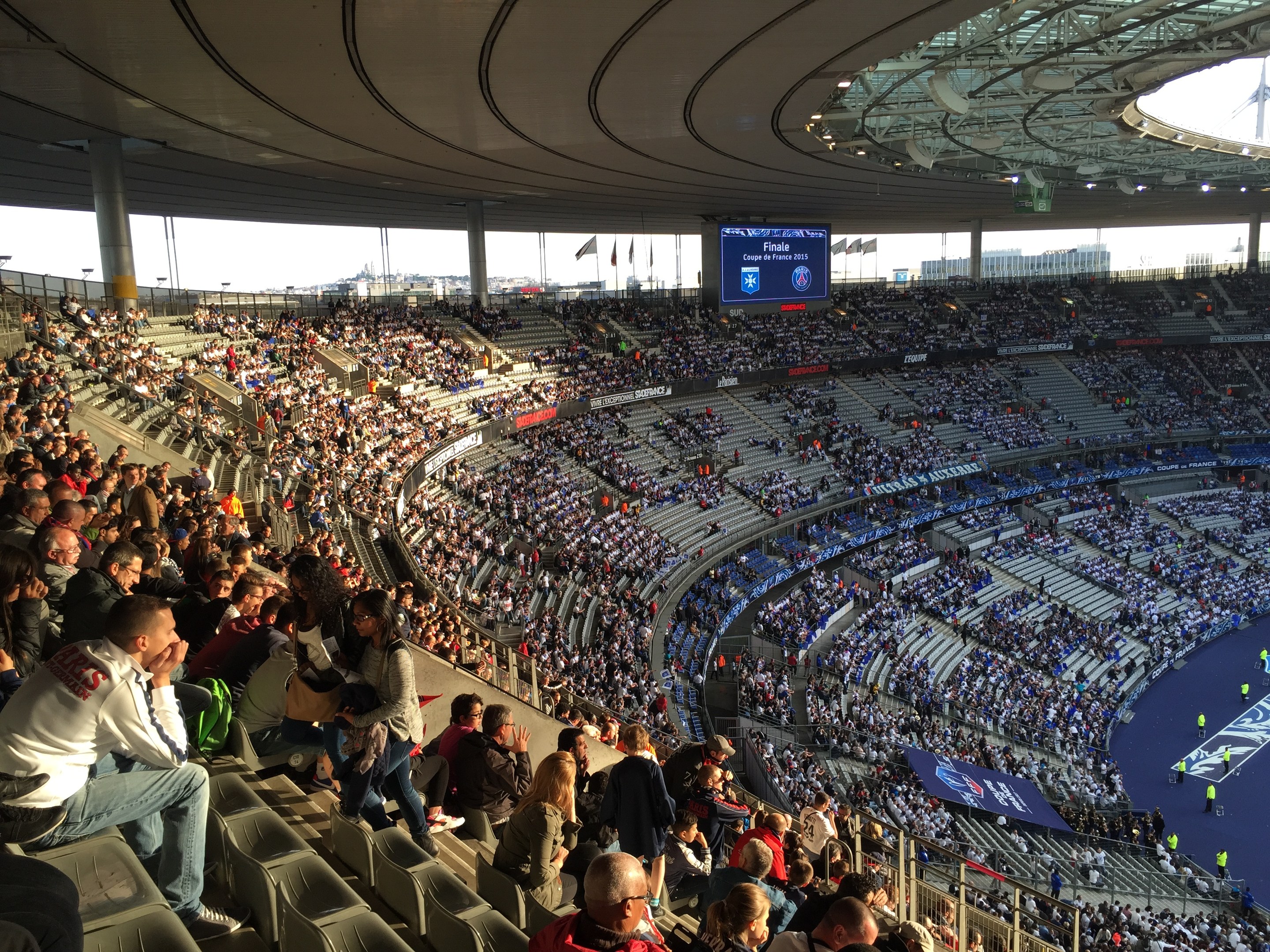 Große Zuschauermenge in einem Stadion bei einem Fußballspiel mit einer Bühne, Fahnen, Stangen, einem Bildschirm und der Allianz Arena in München, Deutschland im Hintergrund.