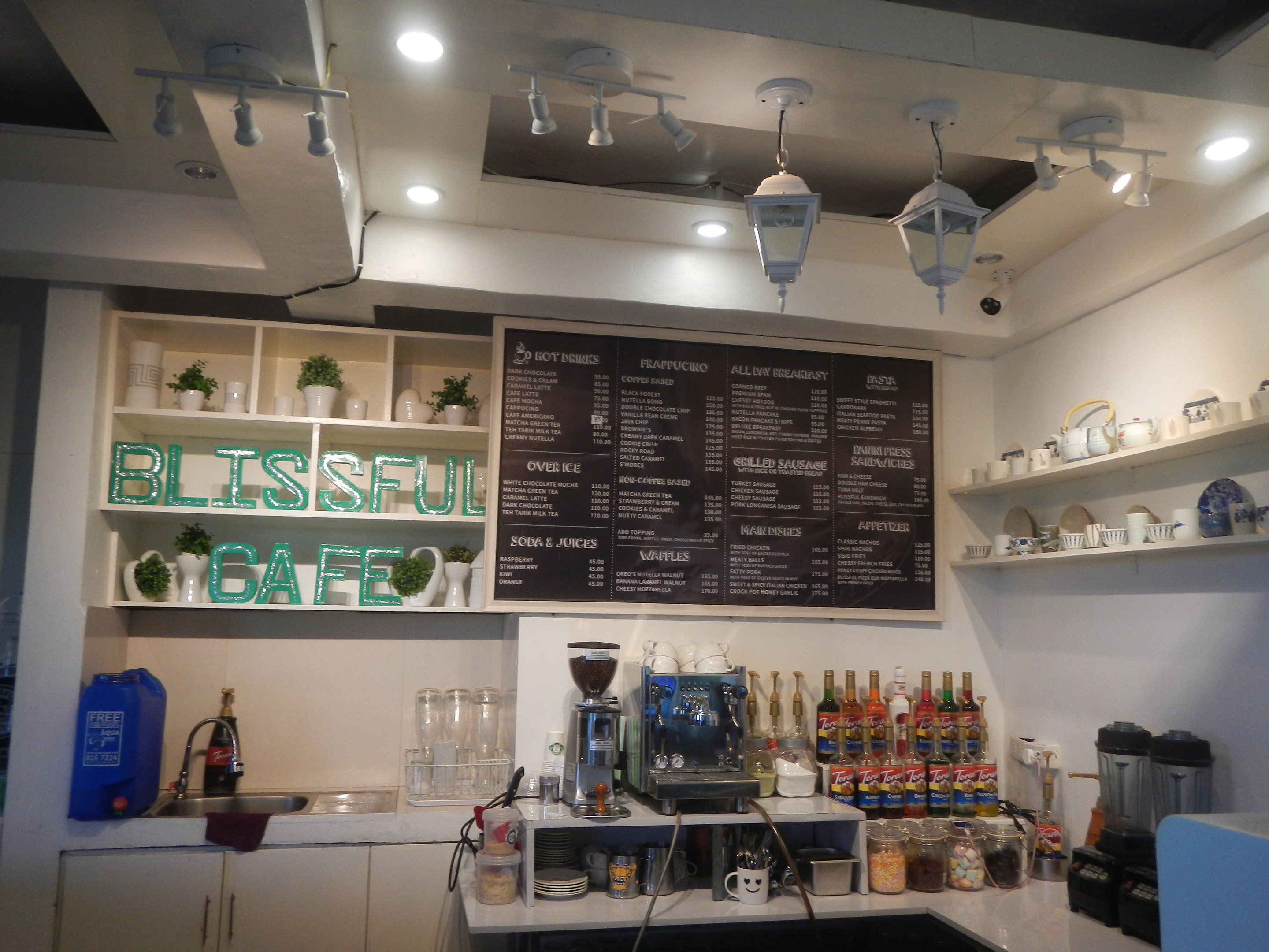 Ein gemütliches Cafe-Interieur mit einer Theke, auf der Flaschen, Tassen und Küchenutensilien, ein Waschbecken links und Regale mit Tellern im Hintergrund unter warmen Deckenlampen und einer Speisekarte.