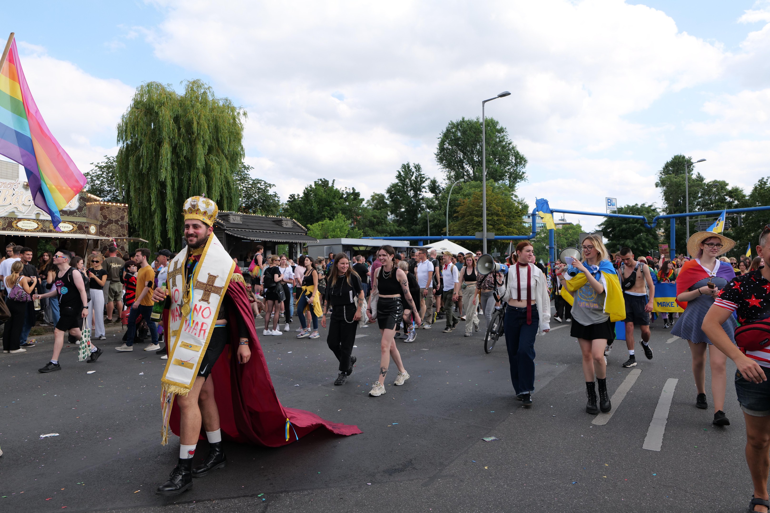 Eine Gruppe von Menschen marschiert auf der Pride Parade 2018 mit einer Regenbogenflagge und Musikinstrumenten, während im Hintergrund Laternenpfähle, Bäume und Hütten zu sehen sind.