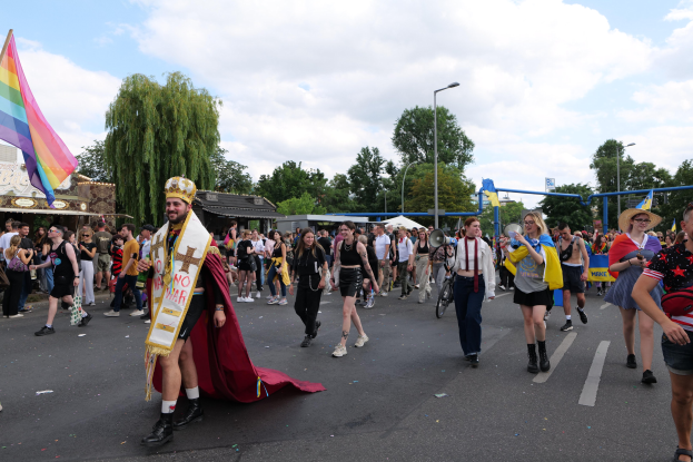 Eine Gruppe von Menschen marschiert auf der Pride Parade 2018 mit einer Regenbogenflagge und Musikinstrumenten, während im Hintergrund Laternenpfähle, Bäume und Hütten zu sehen sind.