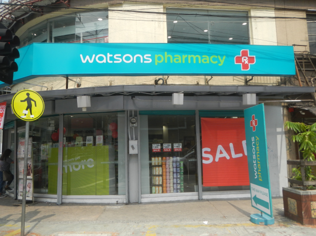 Ein Gebäude mit Glaswänden und -türen, ein Schild mit der Aufschrift "Watsons Apotheke in New York City" auf einem Pfahl, eine Ampel mit einer Lampe, eine Gruppe von Menschen auf dem Gehweg, Topfpflanzen und ein bewölkter Himmel.