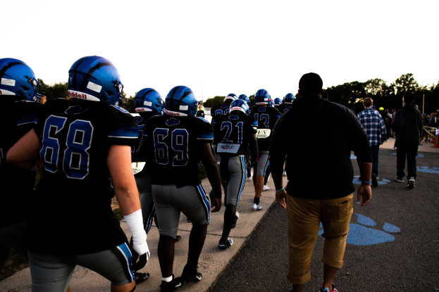 Gruppe von Fussballspielern in Helmen und Stiefeln, die eine Straße mit Bäumen und einem klaren blauen Himmel im Hintergrund entlanggehen.