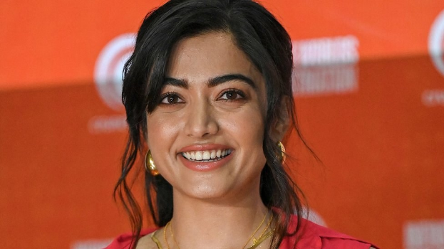 Eine Frau in einem roten Kleid mit einem eleganten Dutt und dezentem Make-up lächelt in die Kamera, trägt schwarze High Heels und hat ein hellgelbes Banner mit der Aufschrift 'Sonam Kapoors beste Beauty-Looks vom Cannes Film Festival' im Hintergrund.