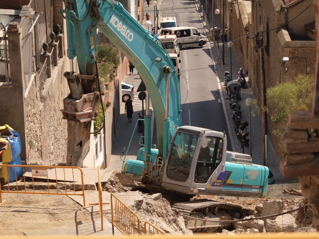 Bagger im Einsatz auf einer urbanen Baustelle mit Gebäuden, Straßeninfrastruktur, Fahrzeugen, Fußgängern und Bäumen.