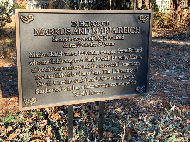 Gedenktafel für Markus und Maria Reich auf einer Tafel mit Text montiert, vor einem Gebäude umgeben von Bäumen und Pflanzen, mit trockenen Blättern auf dem Boden.