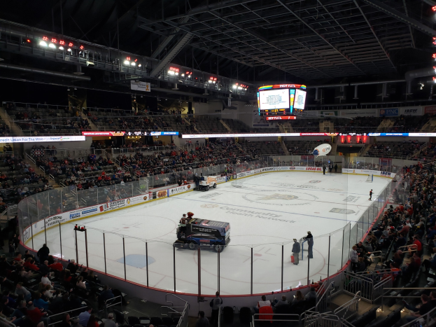 Eishockeyspiel in einer großen Arena mit Fahrzeugen auf dem Eis, Zuschauern in den Rängen und Beleuchtung und Anzeigetafel von oben