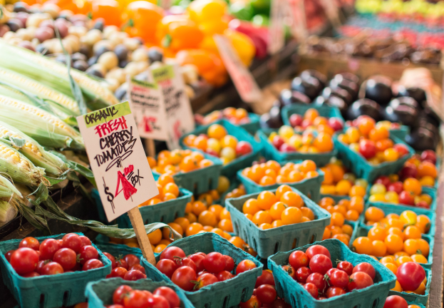Ein Bauernmarkt mit Körben voller frischer Erzeugnisse, darunter Tomaten und Mais, sowie Tafeln mit Text im Hintergrund.