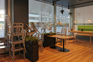 Ein Restaurant mit hölzernen Tischen und Stühlen vor einem großen Fenster, umgeben von Pflanzen und Lichtern, mit Menschen und Gebäuden draußen in Berlin, Deutschland.