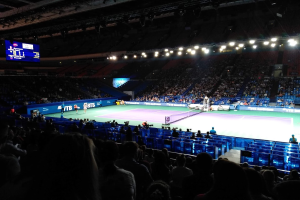 Ein Tennis-Spiel in einer großen Arena mit Zuschauern auf Stühlen, ein Netz teilt den Platz, einige stehende Personen, ein Bildschirm mit Text links und Überdachungsbeleuchtung.