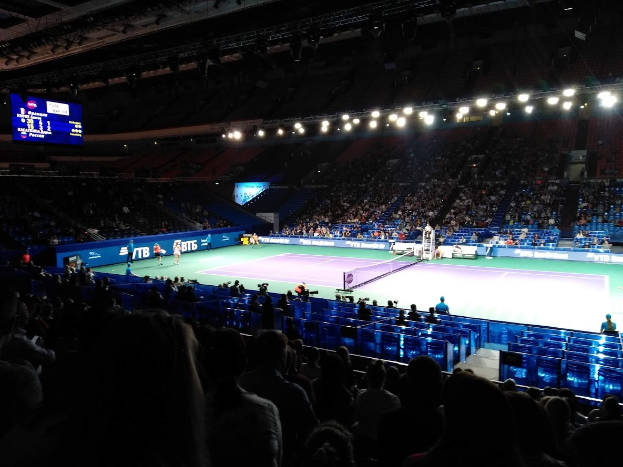 Ein Tennis-Spiel in einer großen Arena mit Zuschauern auf Stühlen, ein Netz teilt den Platz, einige stehende Personen, ein Bildschirm mit Text links und Überdachungsbeleuchtung.