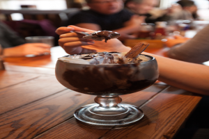 Eine Gruppe von Menschen sitzt um einen Tisch mit einer Schüssel Schokoladenpudding in der Mitte, eine Person im Vordergrund isst mit einem Löffel, Hintergrund leicht unscharf.