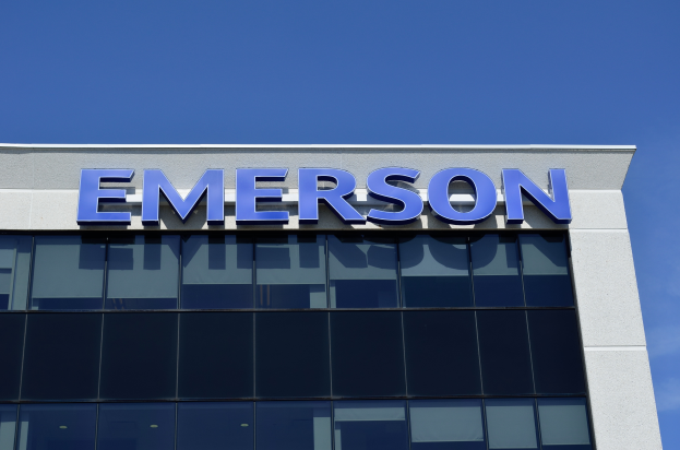 Das Emerson-Logo ist auf der Seite eines Gebäudes mit dem Text "Emerson investiert 1,5 Milliarden USD in den USA" darunter zu sehen, vor einem klaren blauen Himmel.