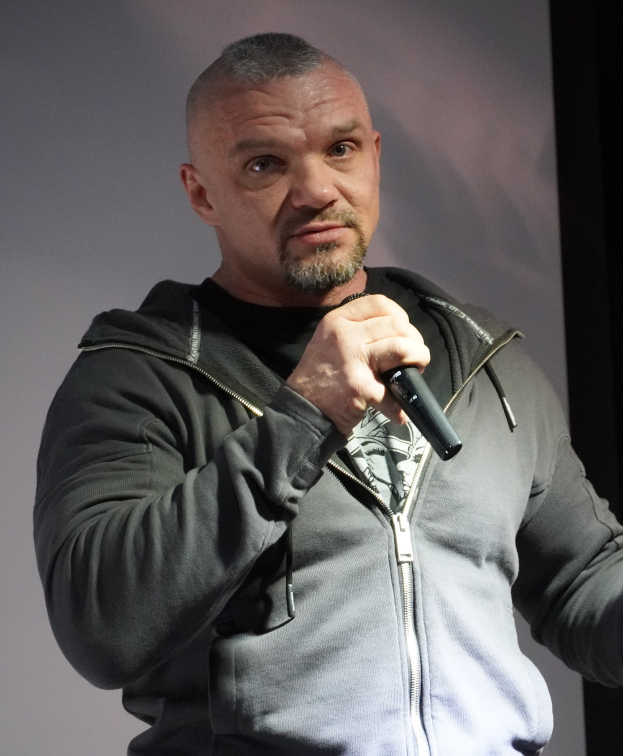 Ein Mann in einer schwarzen Kapuzenjacke und einem schwarzen Anzug mit einem weißen Hemd, der ein Mikrofon hält, steht selbstbewusst mit zurückgekämmtem Haar vor einer Wand.