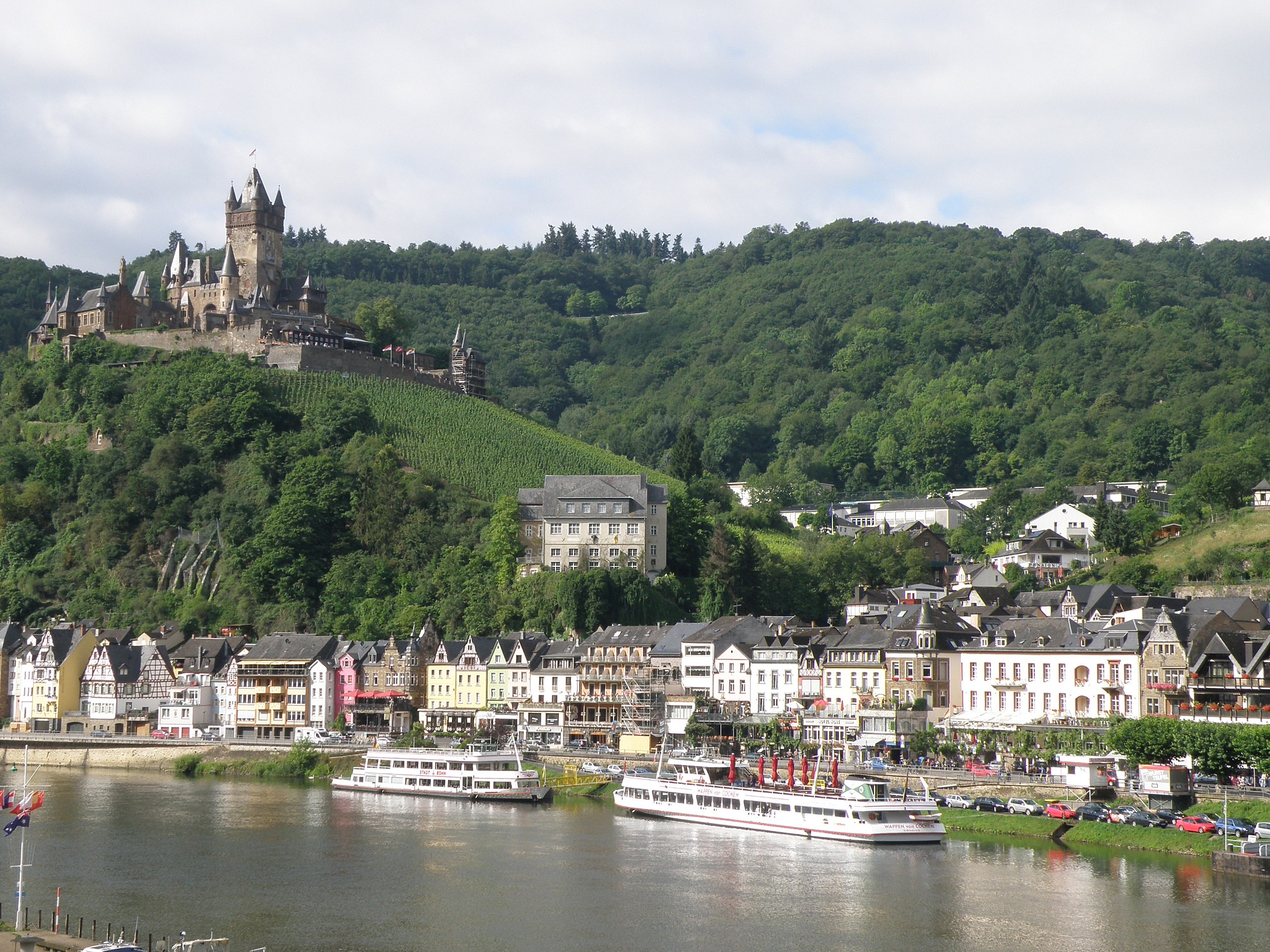 Eine malerische Aussicht auf den Rhein in Deutschland mit einer Burg auf einem Hügel, Booten auf dem Fluss und Fahrzeugen auf der Straße sowie einem bewölktem Himmel.