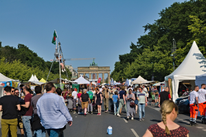 Eine Menschenmenge, die eine Straße entlanggeht, gesäumt von Zelten, Fahrzeugen und Bäumen, mit einem Tor und einem klaren blauen Himmel im Hintergrund und Fahnenmästen auf der linken Seite.