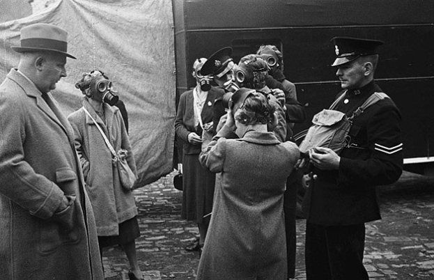 Eine Gruppe von Menschen in Mänteln und Mützen, die Gasmasken tragen, steht zusammen vor einem Zelt, mit einem Fahrzeug im Hintergrund, dargestellt in Schwarz-Weiß.