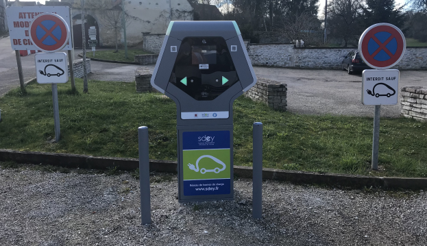 Ein Parkscheinautomat an der Seite einer Straße neben einem Gebäude, umgeben von Gras, Schildern, Fahrzeugen, einer Wand, Bäumen und einem bewölkten Himmel.