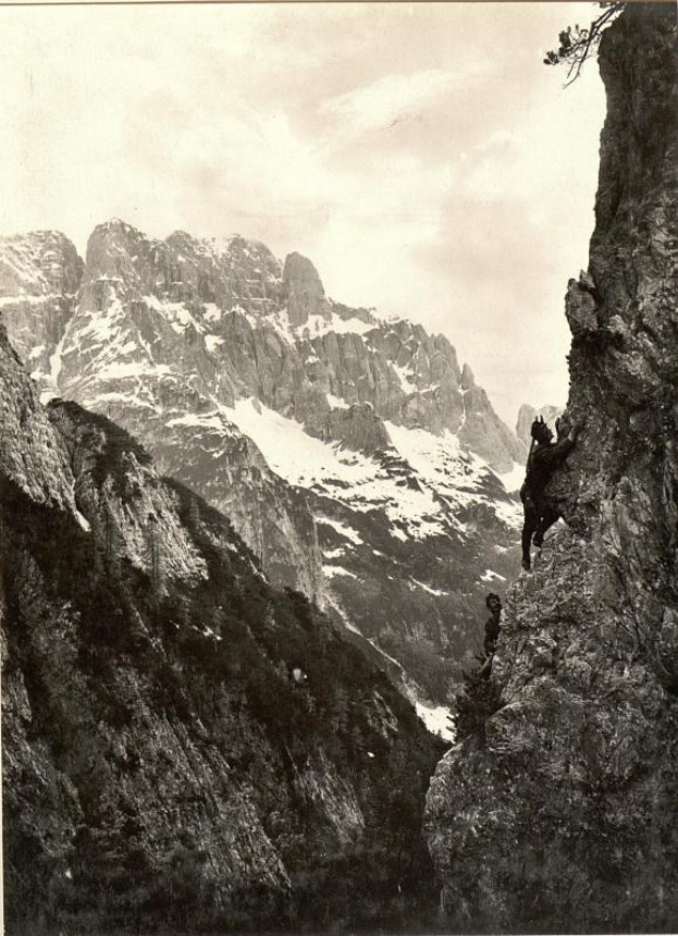 Ein Schwarz-Weiß-Foto eines Mannes, der einen schneebedeckten Berg erklimmt, mit drei prominenten Gipfeln im Hintergrund und dem Text "Gustav Klimt - Die Drei Gipfel" unten.