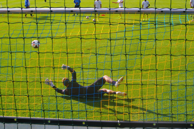 Fußballspieler taucht ab, um einen Schuss mit einem Tor im Vordergrund und Zuschauern im Hintergrund zu halten.
