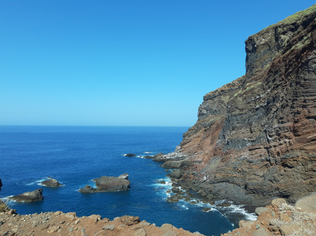 Blick auf das Meer von einer Klippe aus mit Wasser im Vordergrund, Himmel im Hintergrund, einem Hügel auf der rechten Seite und Felsen und Gras unten, der die Landschaft von Lanzarote auf den Kanarischen Inseln zeigt.