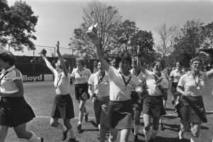Eine Gruppe von Frauen läuft auf einem grünen Fußballfeld, jede hält einen Pokal in der Hand, mit Bäumen, Maschendrahtzaun und Texttafeln im Hintergrund, dargestellt in Schwarz-Weiß.
