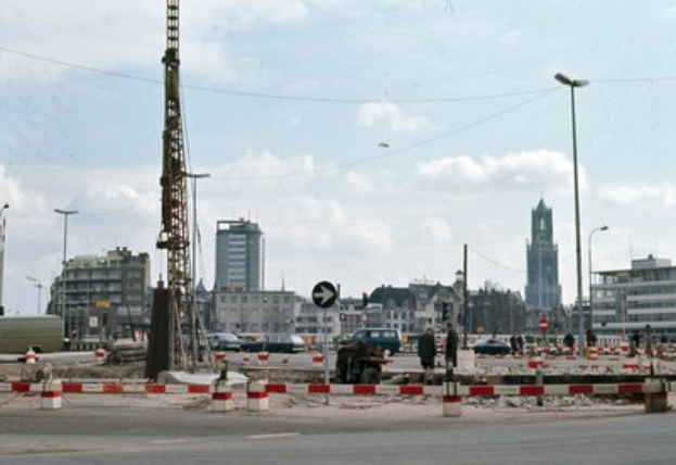 Baustelle mit einem zentralen Turm, umgeben von Gebäuden, Straßeninfrastruktur, Fahrzeugen und Stromleitungen unter einem bewölkten Himmel.